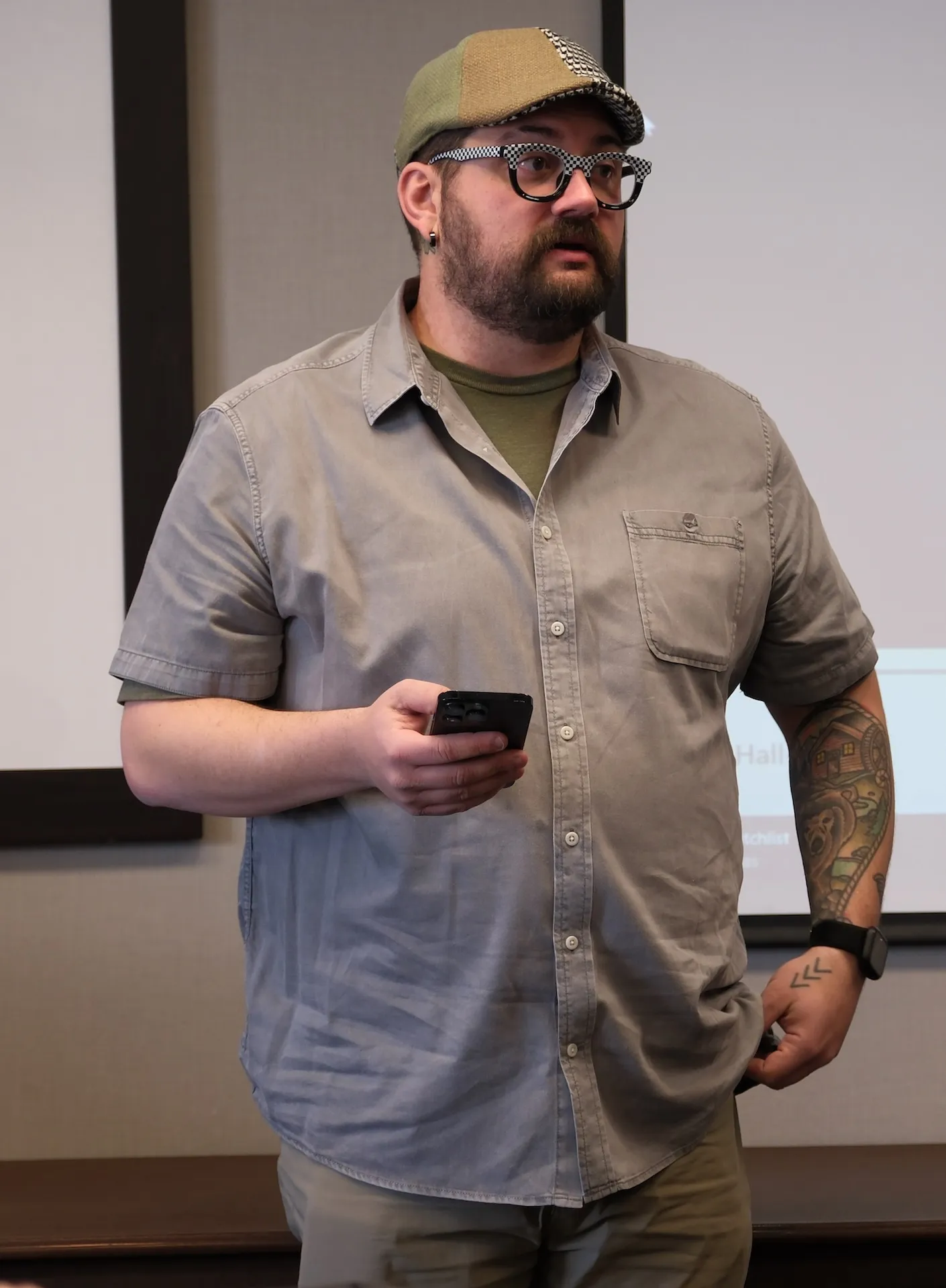 A man with a collared shirt, glasses and flat cap stands in front of room while speaking to group.