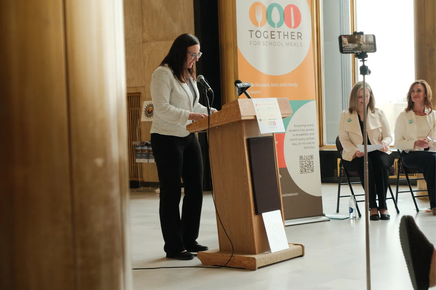 A coalition member speaks at a podium in front of a “Together for School Meals” banner.