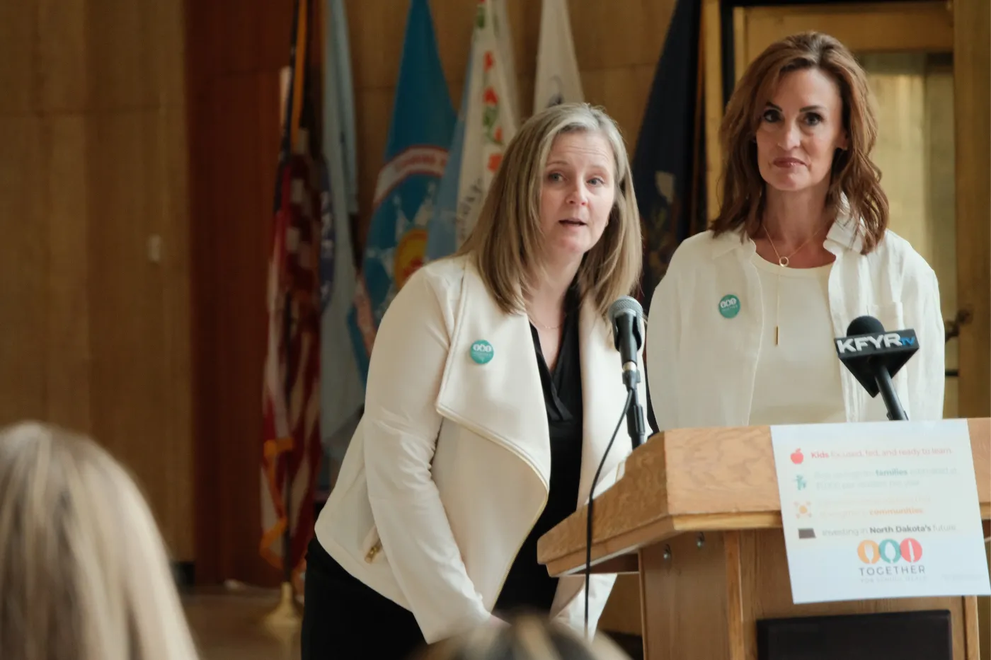 Two speakers stand at a podium addressing attendees during the press event.
