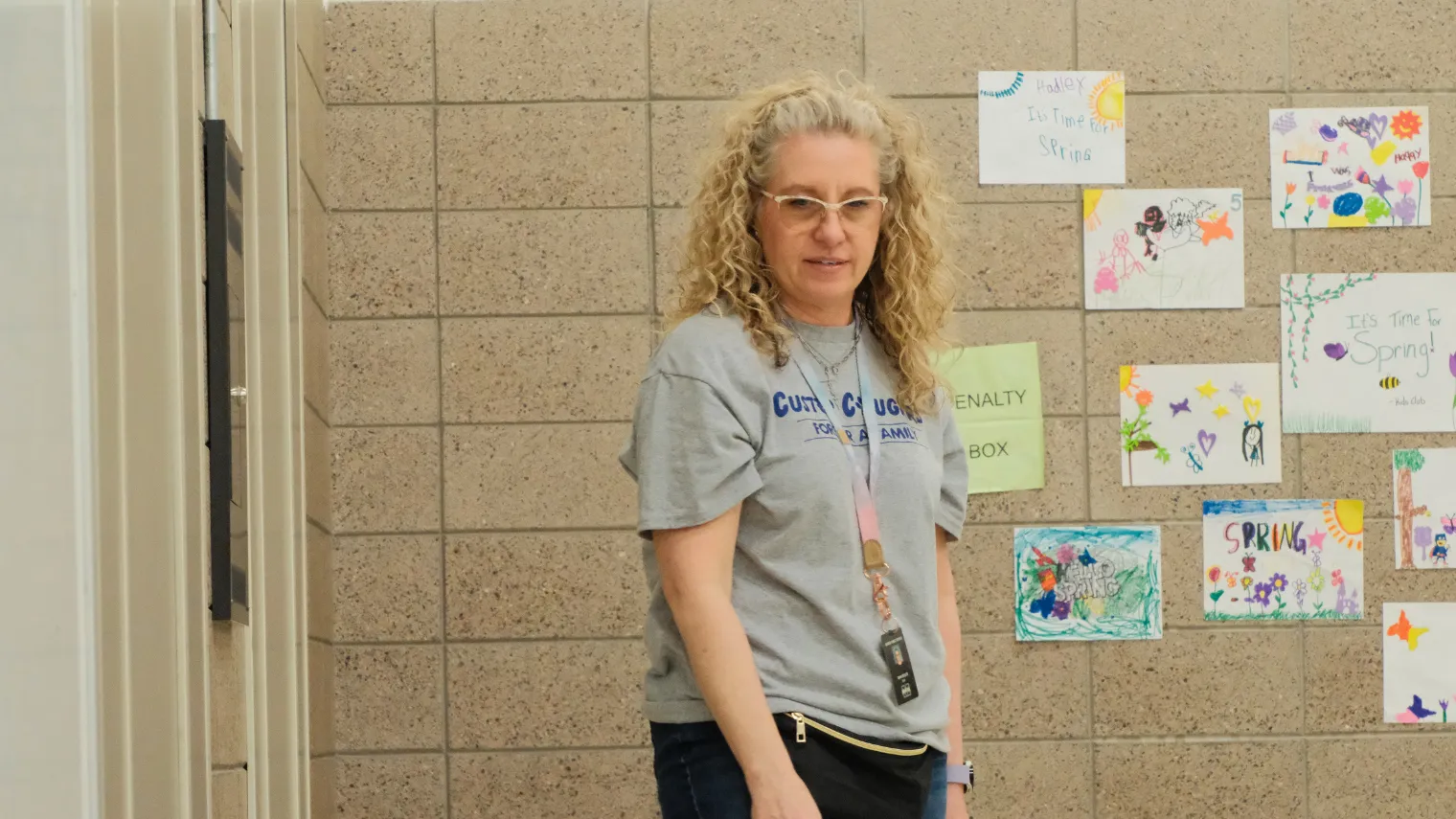 Dawn Ressler stands in a school gym, preparing students for the next activity during a physical therapy session. Children's artwork is displayed on the wall behind her.