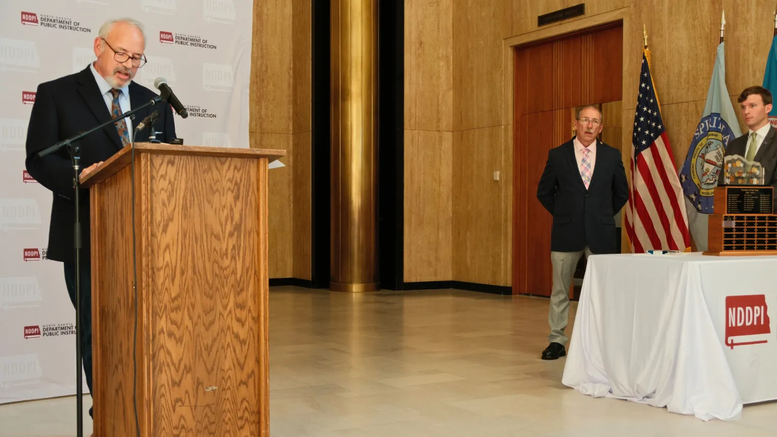 North Dakota United President Nick Archuleta speaks from behind a podium while 2027 ND Teacher of the Year Brian Schneider and State Superintendent Levi Bachmeier stand off to the side.
