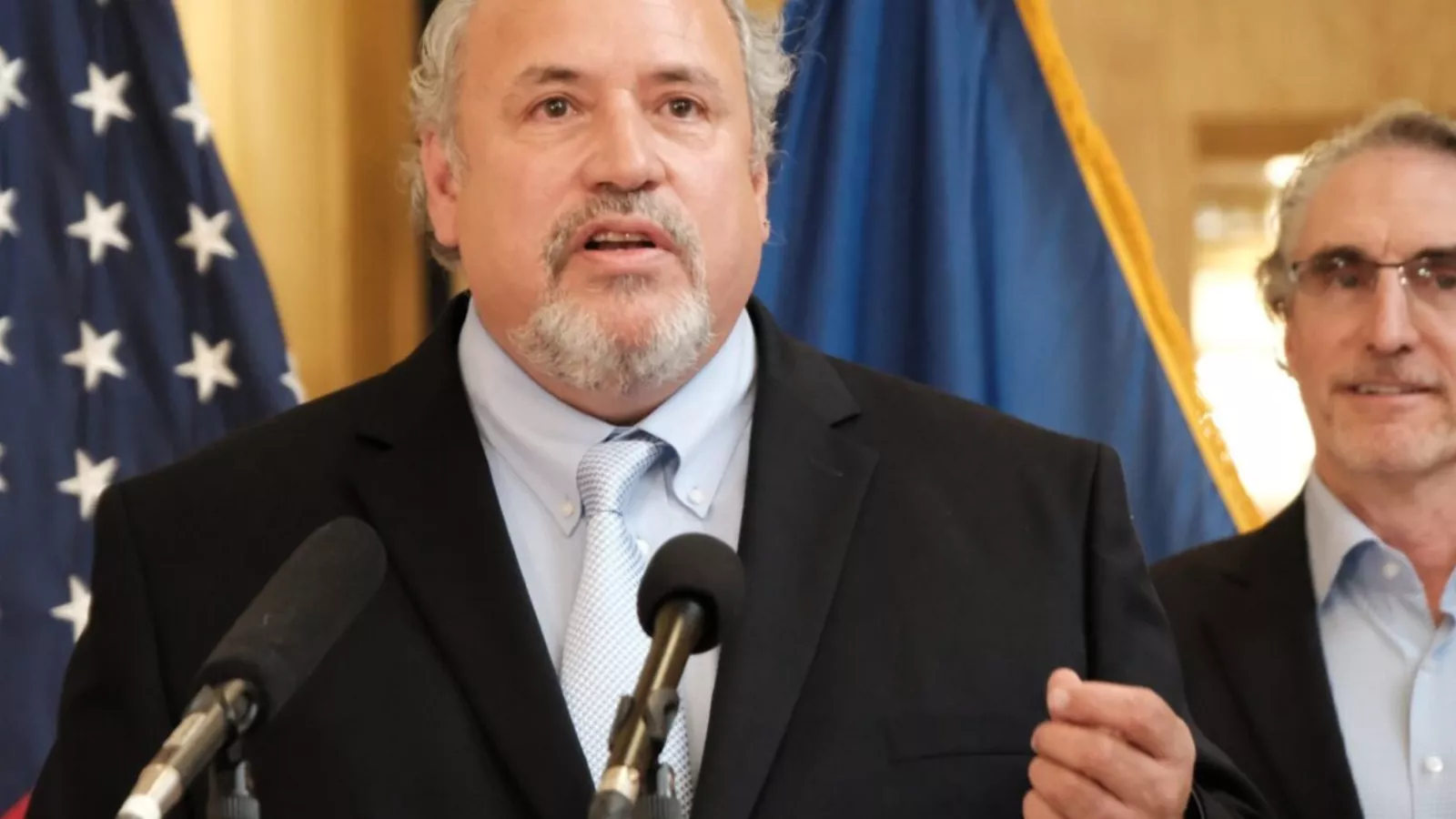North Dakota United President	Nick Archuleta speaks into microphones at a press conference. Behind him there is a glimpse of the American flag and the North Dakota flag.