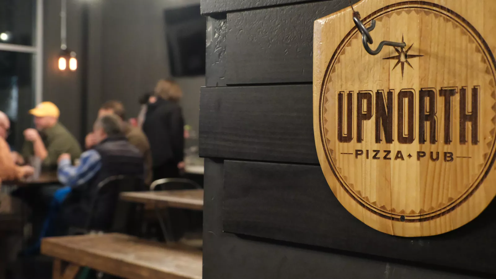 Group of people gathered around table in background, behind sign that reads "Up North Pizza & Pub."