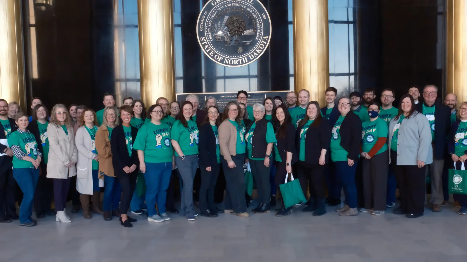A large group of people — some wearing green T-shirts printed with "Lobby Day" and the ND United logo — stand together inside Memorial Hall of the state Capitol building.