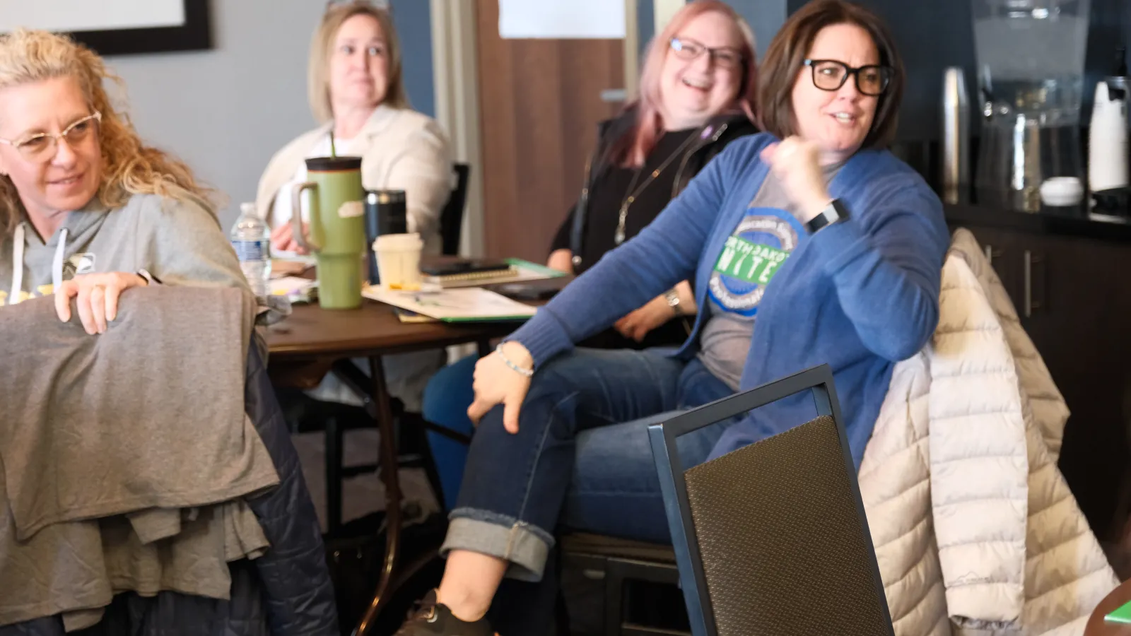 A group of four women sit around a table in a conference room, all looking backwards at a speaker who is outside of frame.
