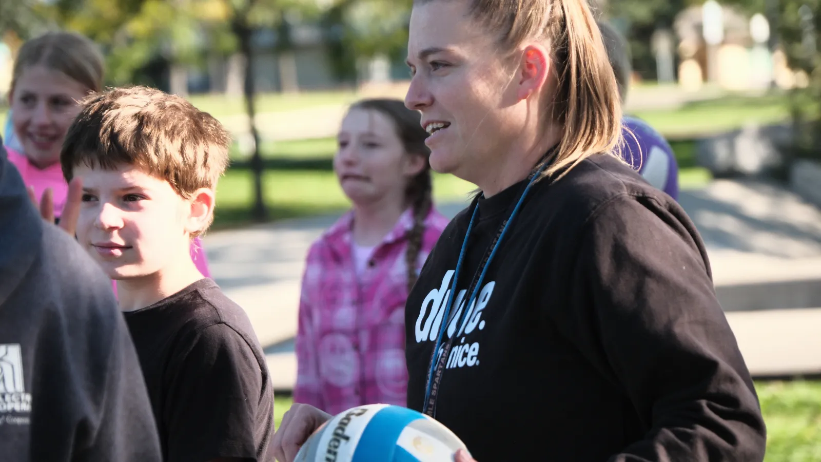 A teacher holds a volleyball while standing with students outdoors, giving instructions during a physical education activity on a sunny day.