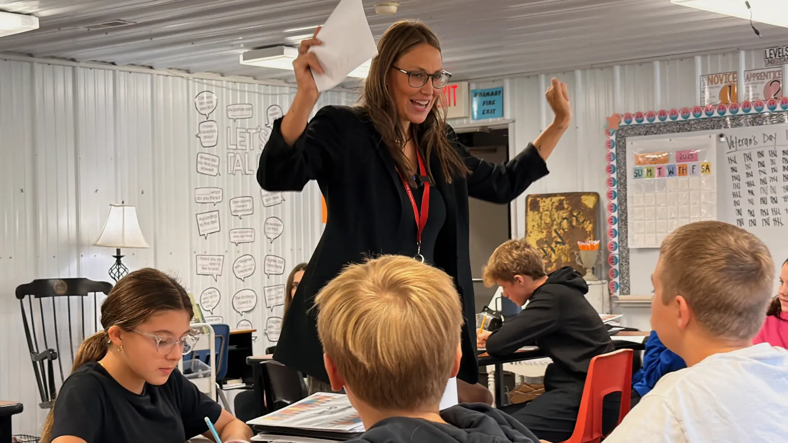 A teacher stands beside a group of students seated at desks, raising both hands while holding a sheet of paper as she speaks. Students around her are writing and listening during a classroom lesson.