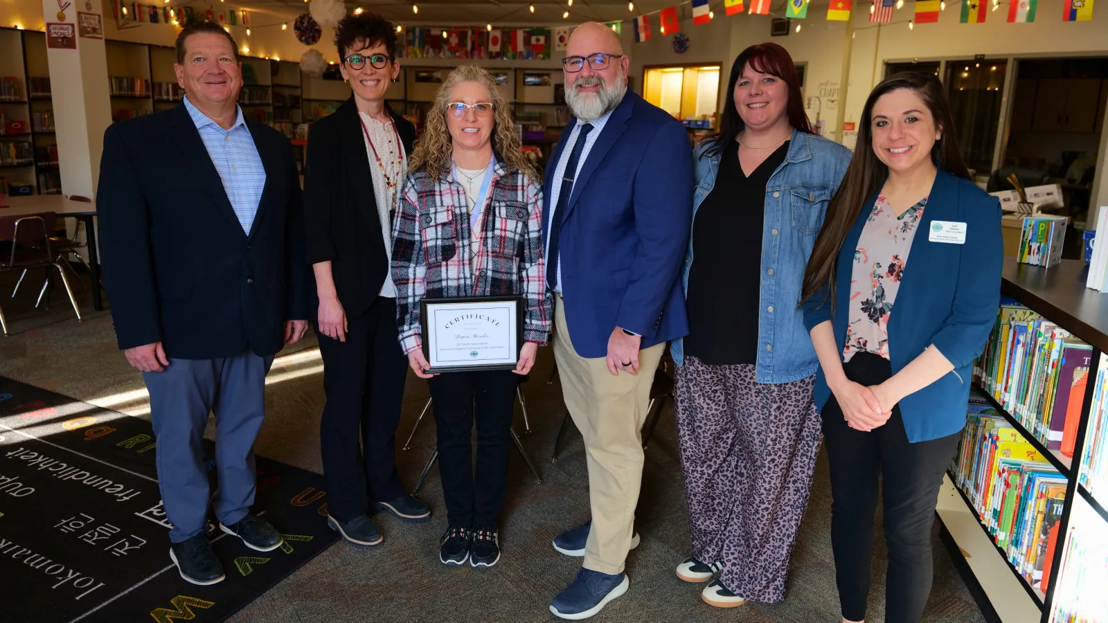 A group of six adults stand together in a school library, with one woman holding a framed certificate while colleagues and administrators gather around her in celebration.