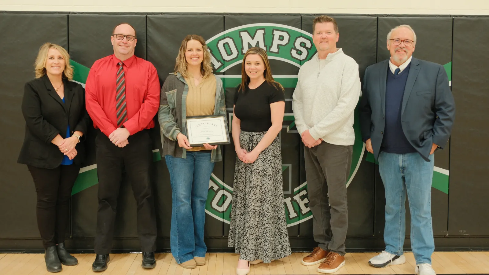 Six adults pose in a school gymnasium in front of a “Thompson” wall graphic, with one woman at center holding a certificate recognizing her achievement.