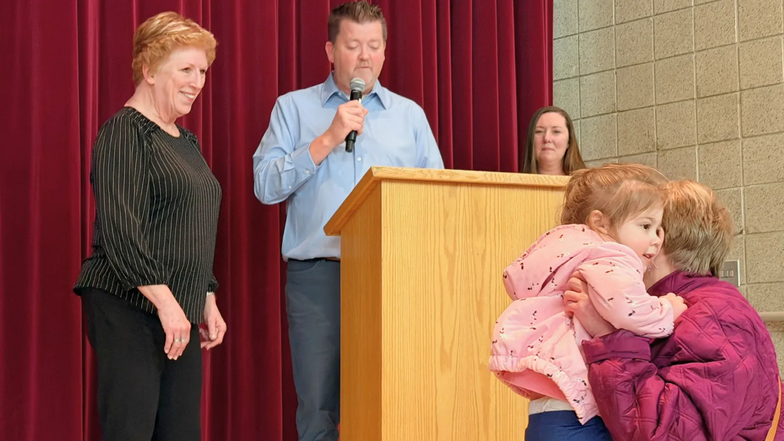 At a school assembly, a man speaks at a podium on stage while a woman nearby smiles and another adult holds a young child in the foreground, capturing a candid moment during the event.