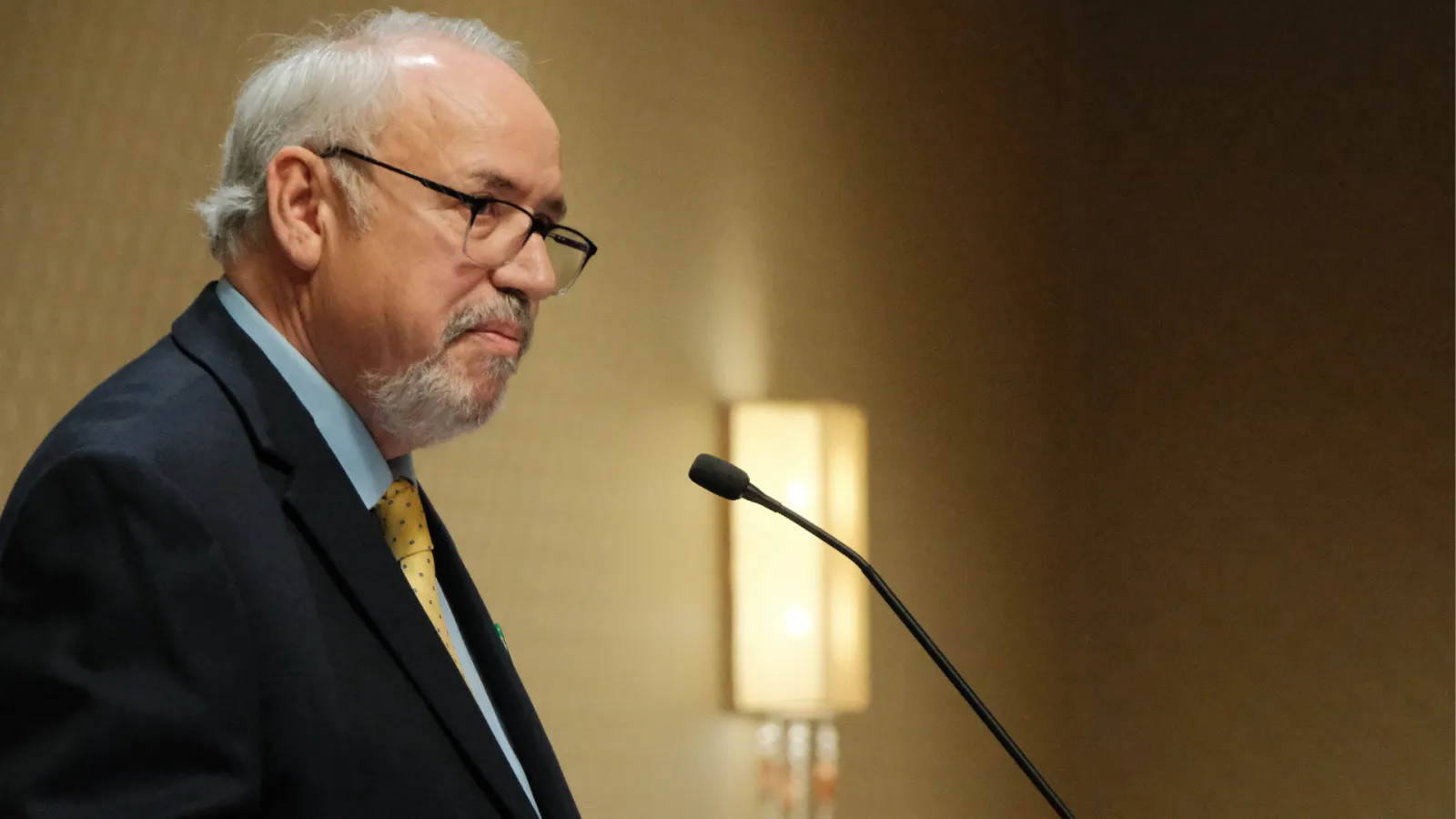 North Dakota United President Nick Archuleta speaks at a podium during the 2026 North Dakota United Delegate Assembly in Bismarck. He is shown in profile wearing a dark suit, light blue shirt and gold tie, speaking into a microphone.