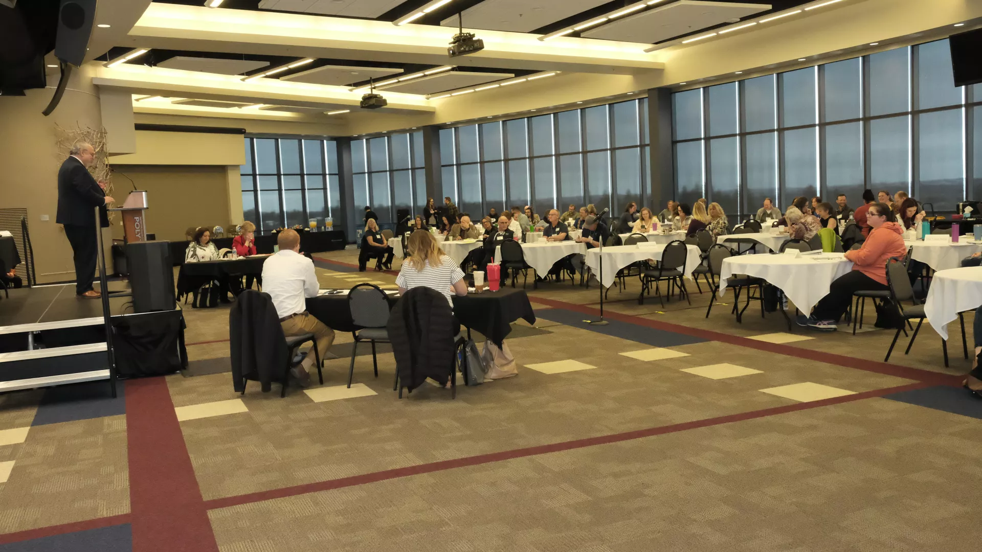 Delegates from across the state of North Dakota, including educators and public employees, sit around multiple tables in a large room listening to North Dakota United President Nick Archuleta speaking.