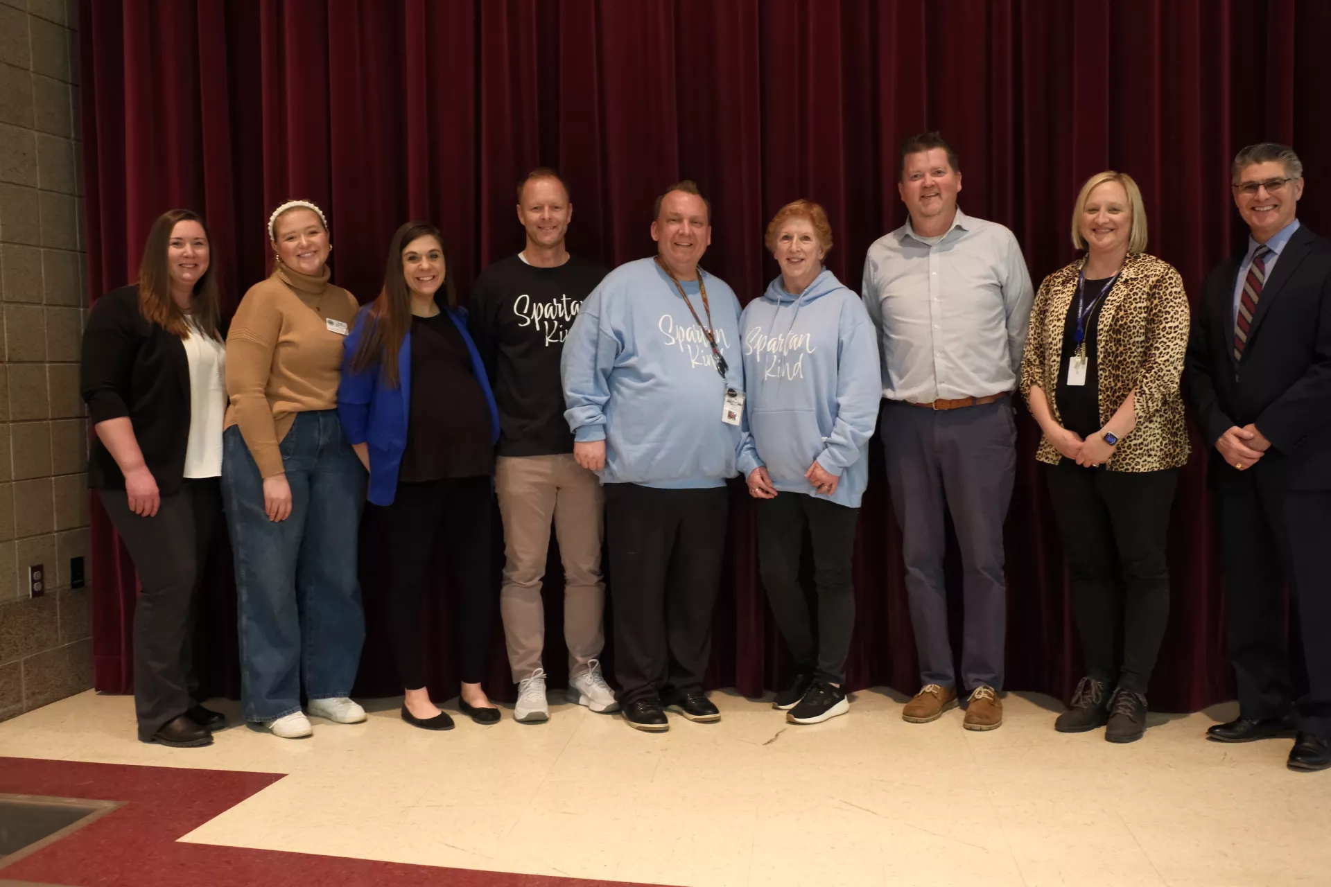 A group of nine people standing in a row in front of a red curtain. They are dressed in a mix of casual and formal attire, with some wearing hoodies and others in blazers. They are all smiling at the camera.