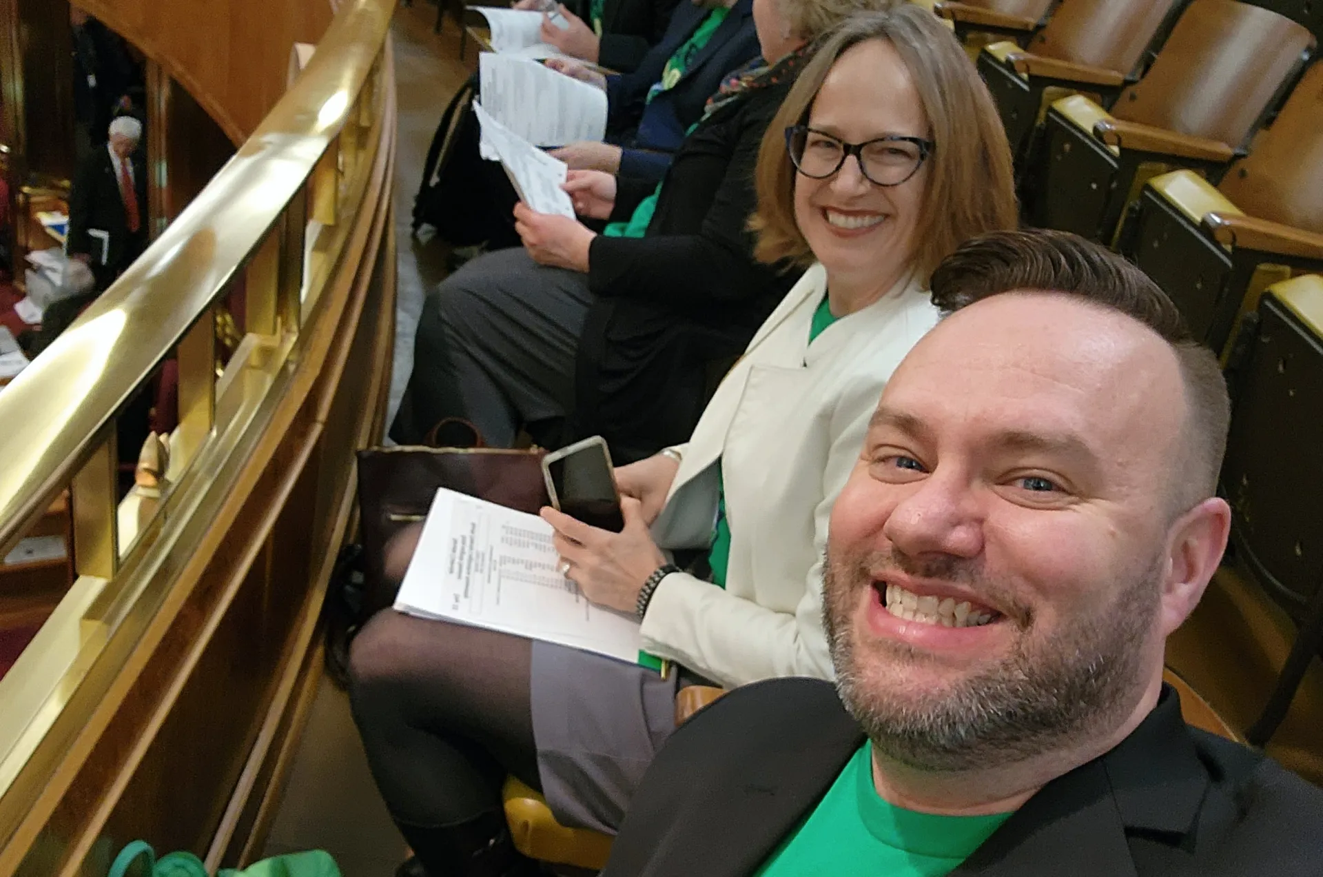 A man in green shirt and black blazer and woman wearing glasses and holding a sheet of paper and cell phone, sitting in a balcony behind a brass railing.
