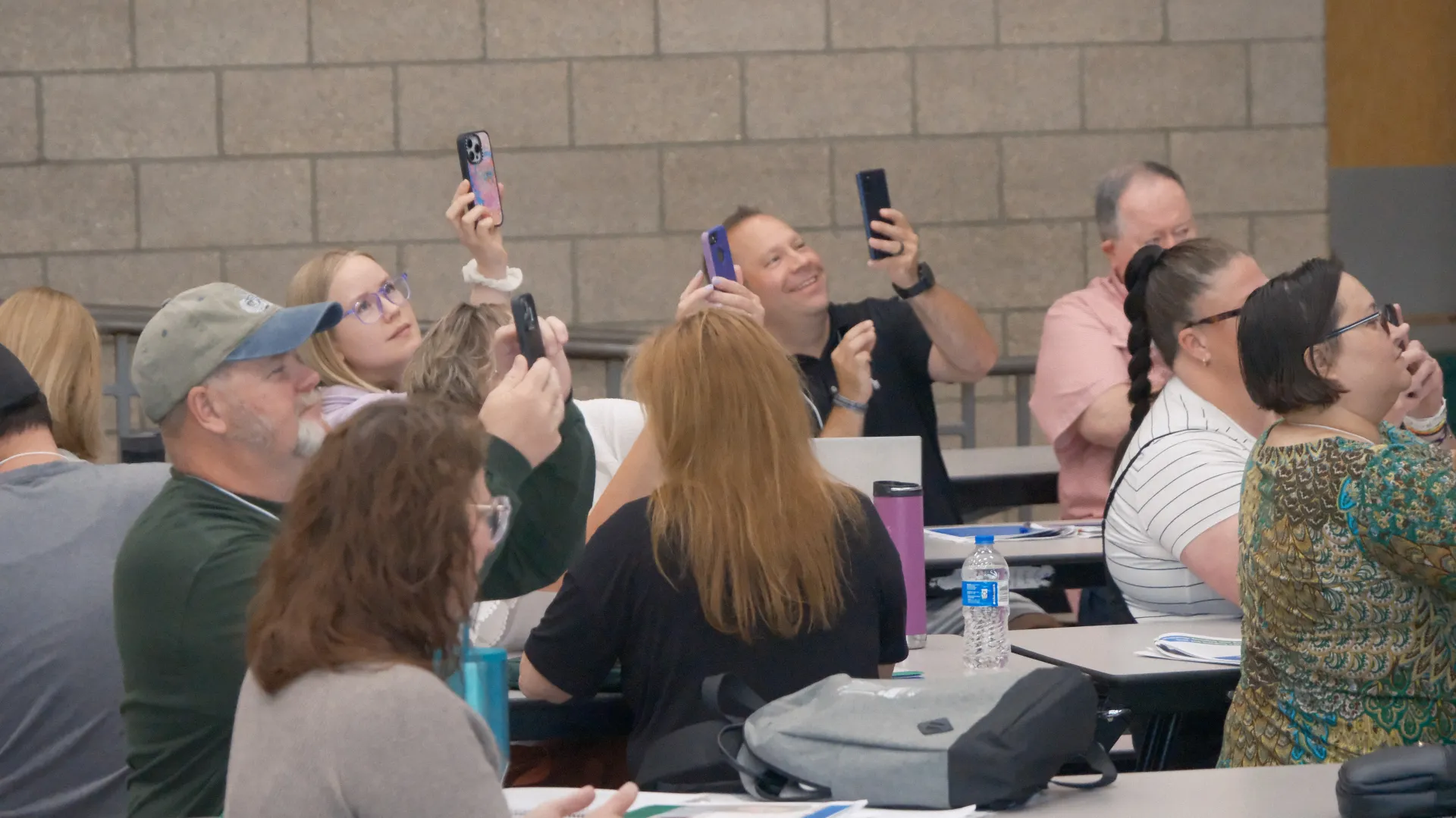 Group of people seated at tables in school cafeteria, raising their mobile phones in air. 