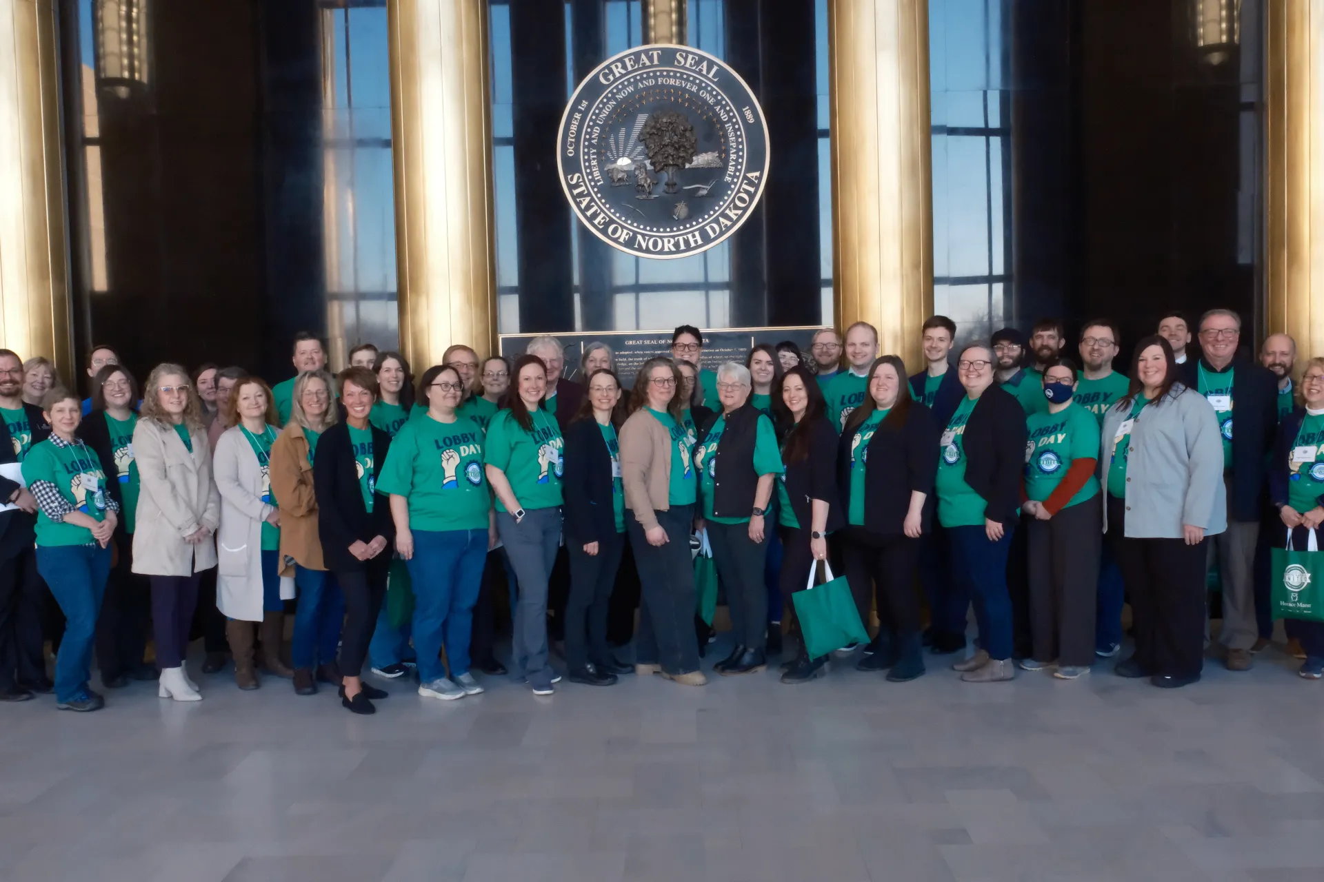 A large group of people — some wearing green T-shirts printed with "Lobby Day" and the ND United logo — stand together inside Memorial Hall of the state Capitol building.