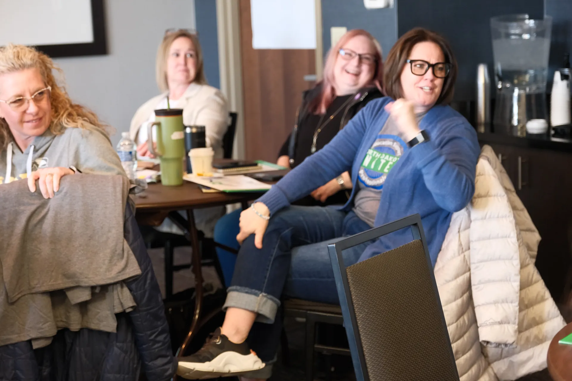 A group of four women sit around a table in a conference room, all looking backwards at a speaker who is outside of frame.
