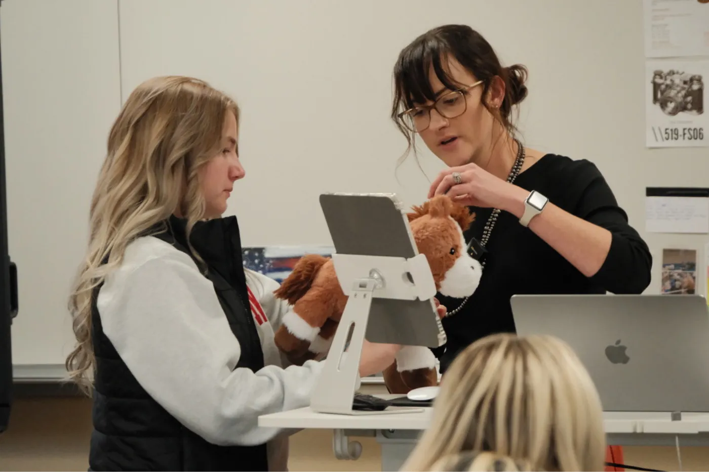 A teacher and a student stand together at a podium, adjusting a plush horse toy in front of a tablet while discussing or demonstrating a project, with other students nearby.