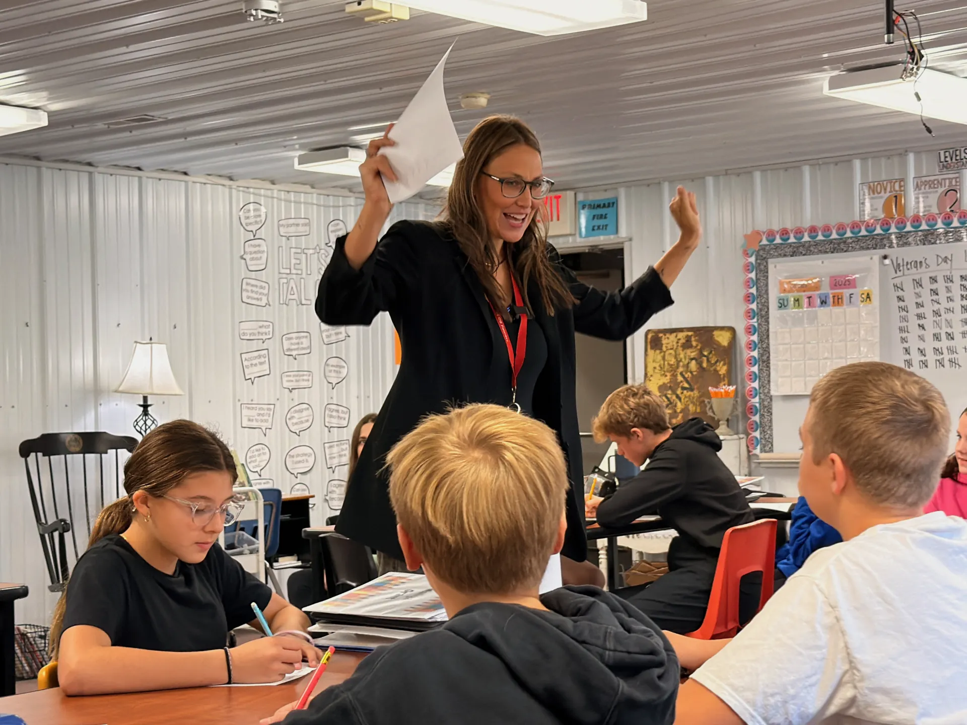 A teacher stands beside a group of students seated at desks, raising both hands while holding a sheet of paper as she speaks. Students around her are writing and listening during a classroom lesson.