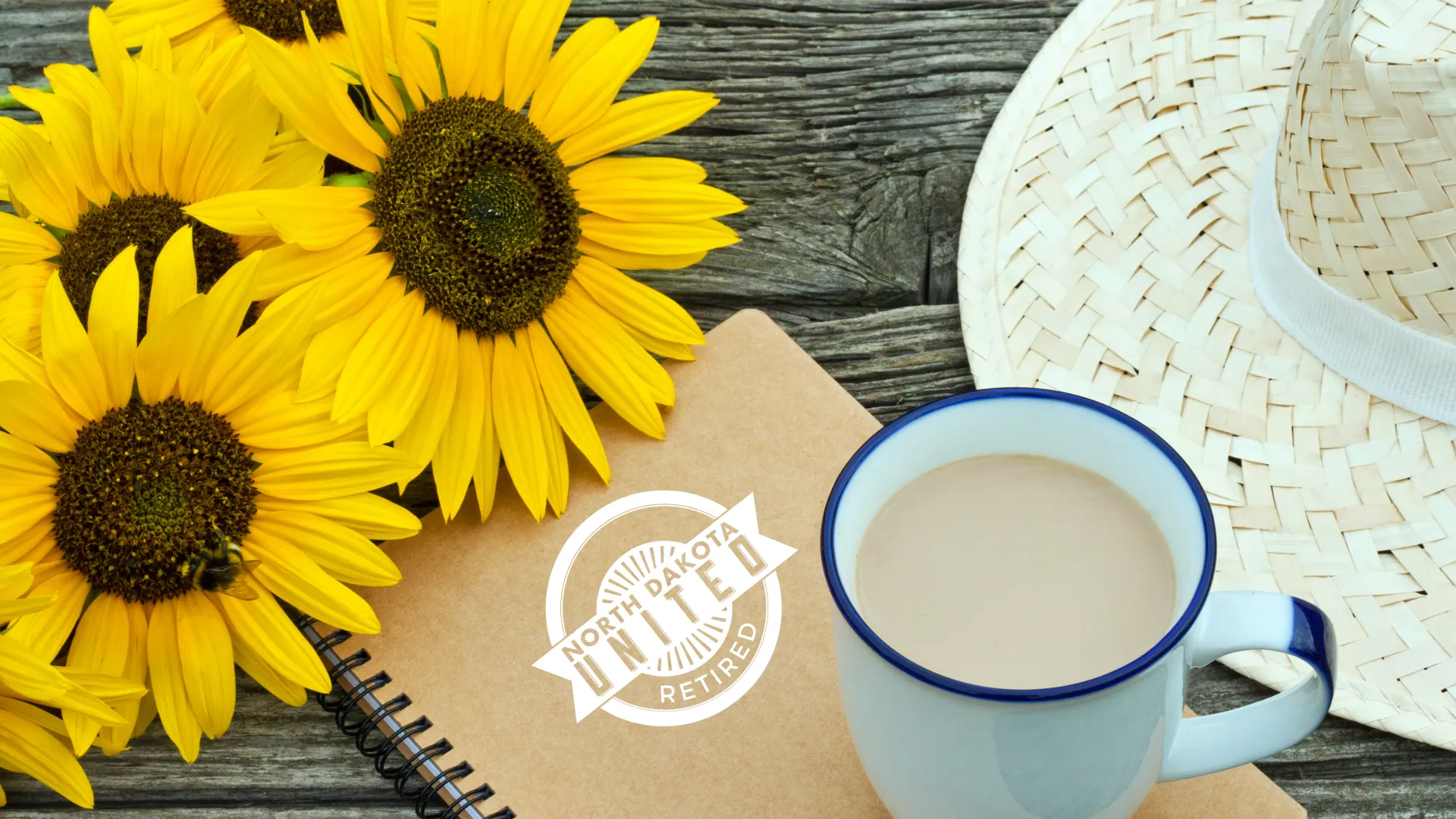 Close-up on three yellow sunflowers, a notebook with North Dakota United-Retired written on the cover, a mug of coffee and a sunhat, sitting on top of a wooden picnic table.