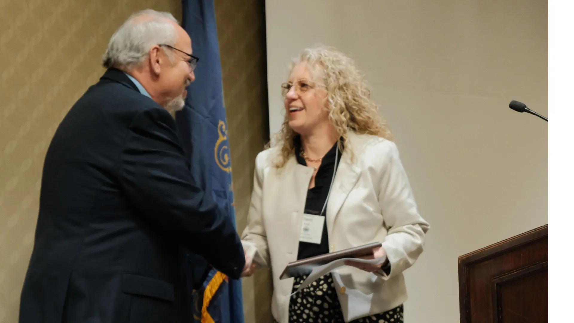 A woman holding a certificate smiles as she shakes hands with a man during an award presentation beside a podium and state flag.