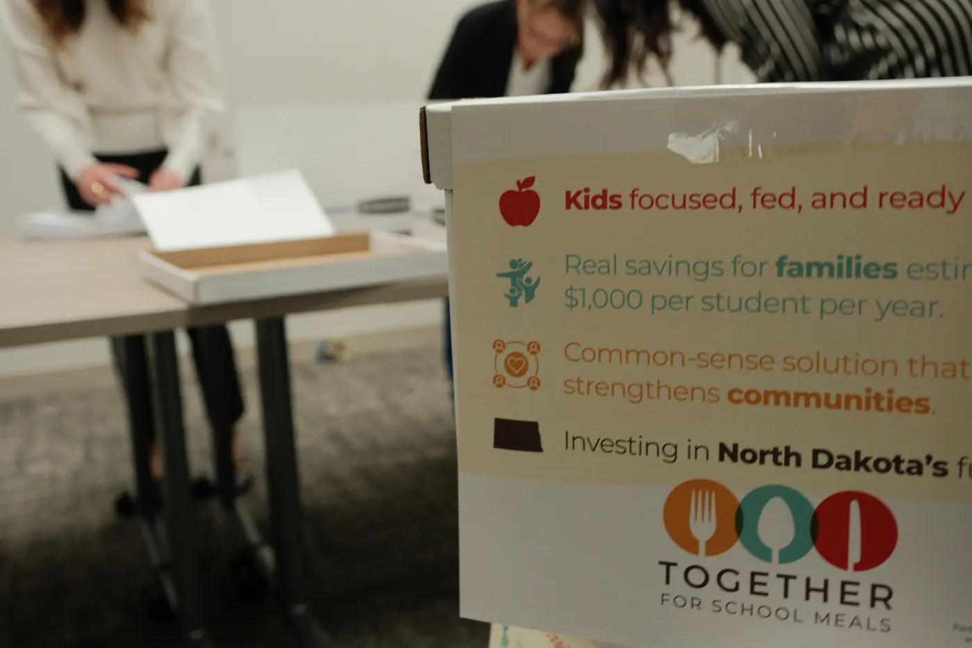 A box labeled with campaign messaging sits in the foreground as Secretary of State staff sort petition sheets in the background.