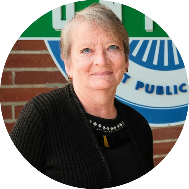 North Dakota United-Retired President Toni Gumeringer standing in front of NDU logo on brick wall in Bismarck. She is shown wearing a black sweater and shirt, smiling at camera.