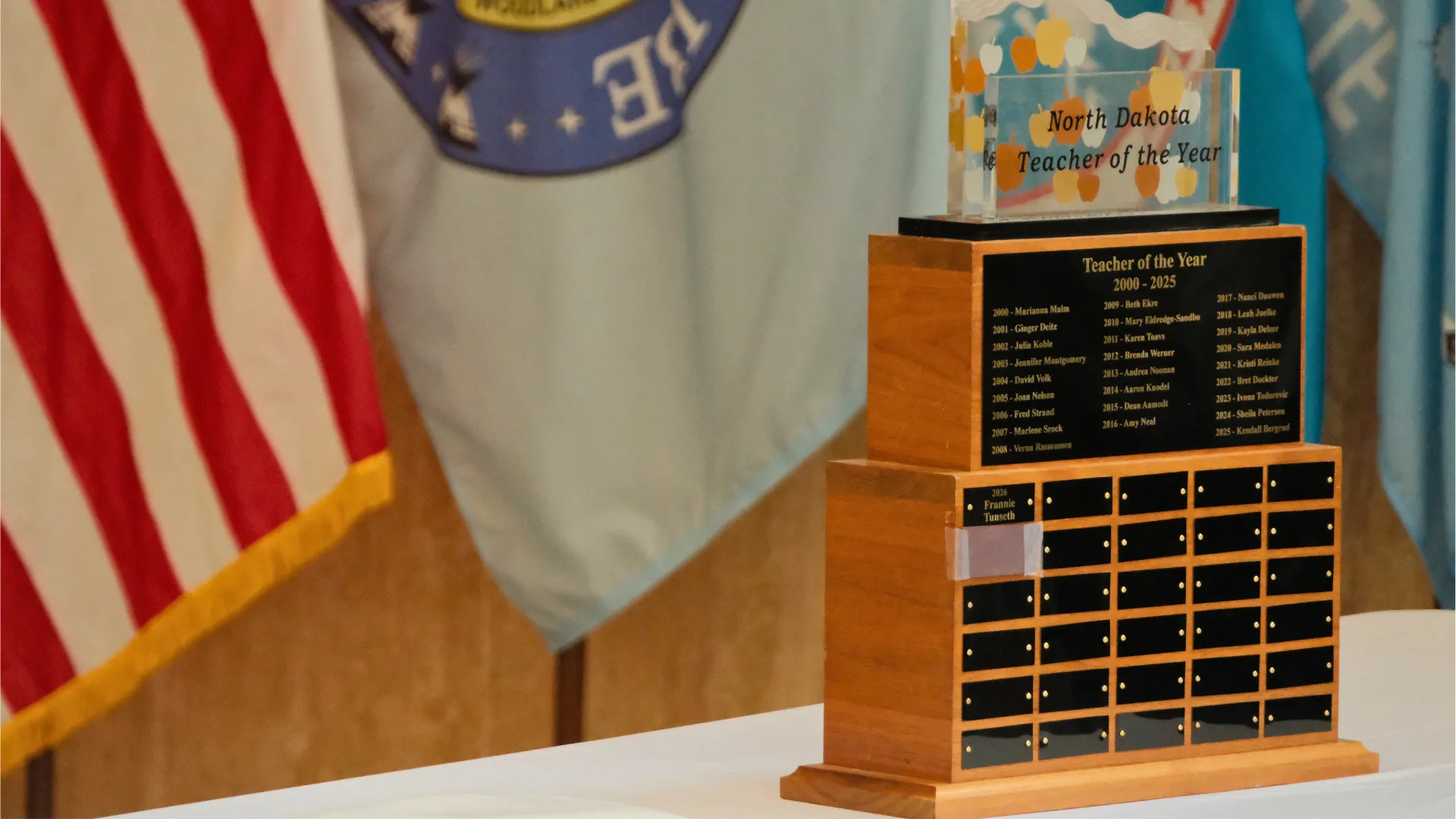 North Dakota Teacher of the Year trophy — listing all winners going back to 1963 — sits on table with with tablecloth in state Capitol with flags in background.
