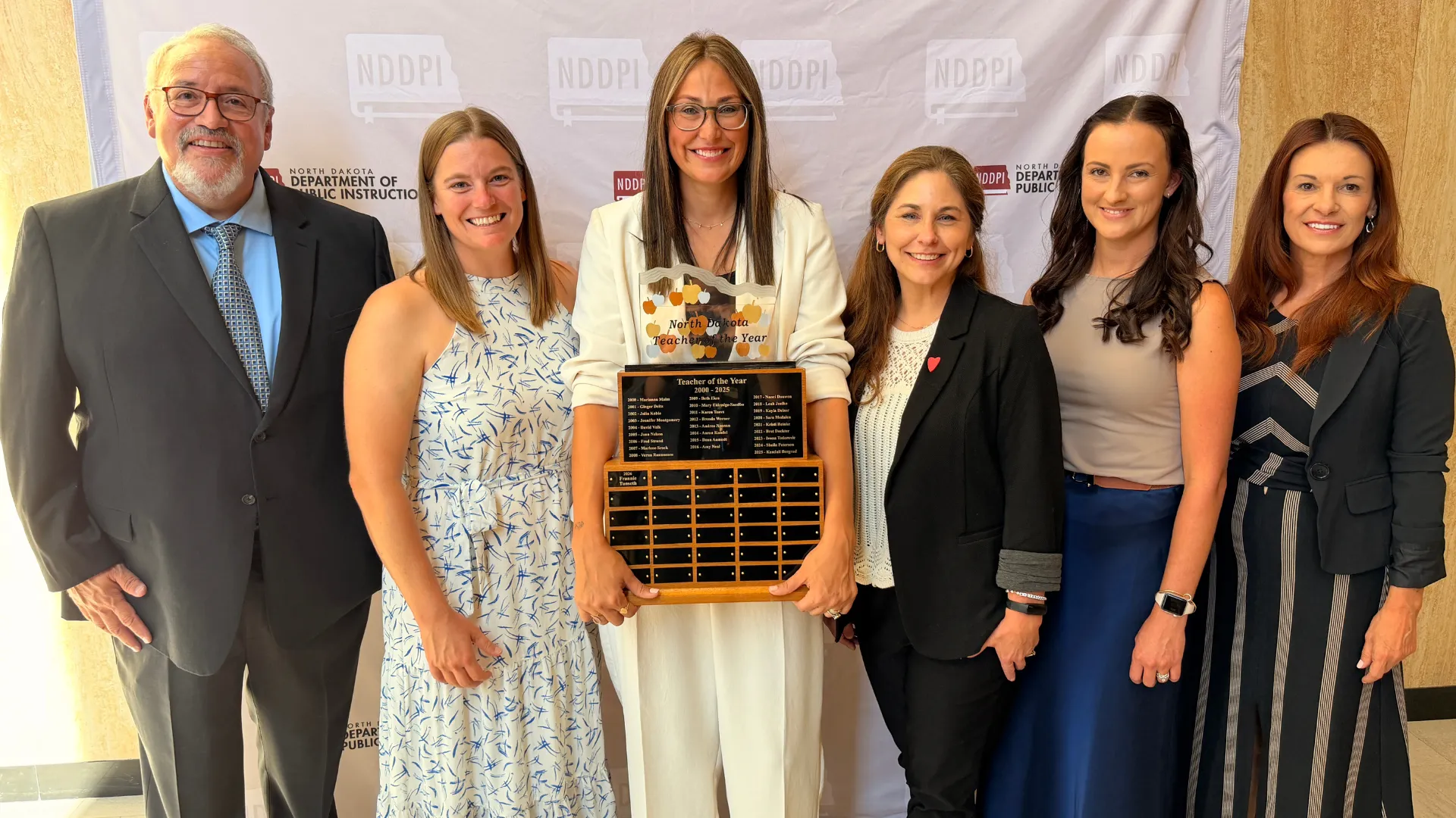 Six individuals standing in a row, posing for photo, with woman in middle holding North Dakota Teacher of the Year trophy.