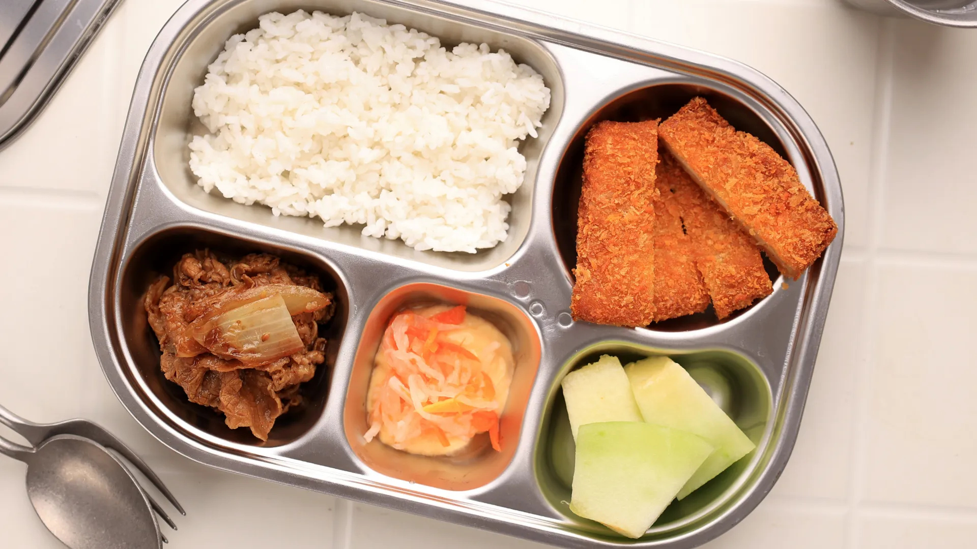A gray, plastic tray of school meal foods in five different compartments, on top of a white tabletop and silver eating utinself surrounding.
