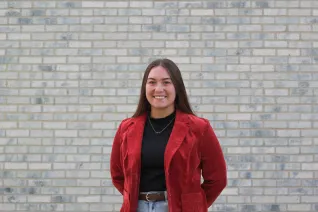 Young woman in red blazer, standing in front of white brick wall.