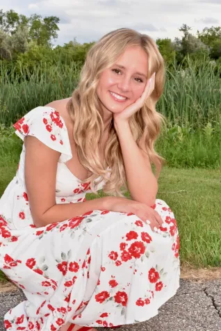Young woman wearing a white dress imprinted with red flowers, crouched down in an outdoor setting.