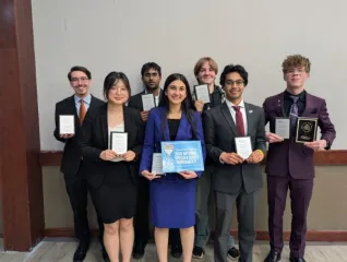 Six students and their advisor stand in front of a wall, holding awards.