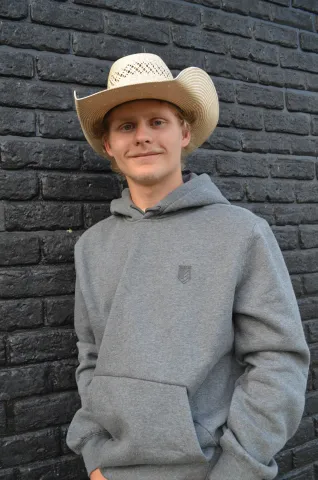 Young man wearing grey sweatshirt and white cowboy hat leans against brick wall.