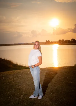 Young woman in white shirt and blue jeans, standing in front of lake as sun sets.