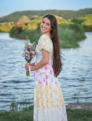 Young woman in multi-patterned dress with long hair, holding flowers and standing near body of water