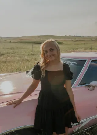 Young woman wearing black dress, standing next to pink automobile