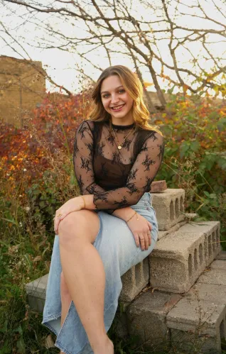Young woman with shoulder-length hair, sitting on cinder blocks outdoors with tree in background