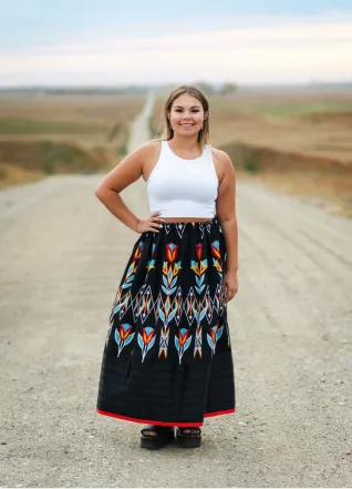 Young woman in white top and black, beaded skirt, standing on gravel road