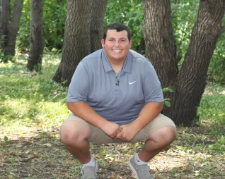 Young man with short hair in shorts and light blue polo shirt, squatted down in front of row of trees.