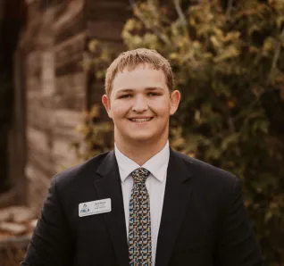 Young man in black suit with white collared shirt and checkered tie, standing outside in front of cabin