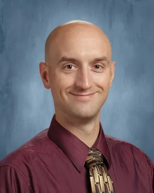 Man in maroon-colored dress shirt and tie, in front of blue background.