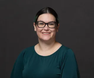 Young woman in glasses and green shirt, in front of dark grey background.