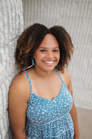 Young woman in light-blue shirt, standing in front of white, patterned wall.