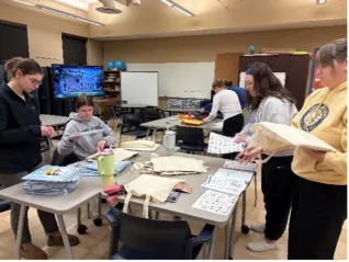 Group of students stand around tables in a classroom, sorting and assembling stacks of small tote bags and paper materials.