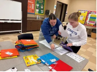 Two students work together at a table, organizing papers and colorful reusable bags laid out in neat piles.