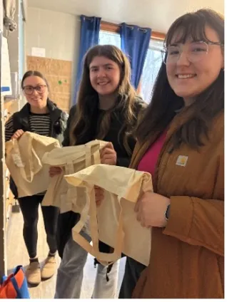 Three students smile while holding plain canvas tote bags inside a classroom with sunlight streaming through the window.