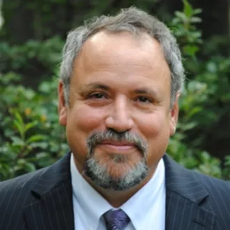 Nick Archuleta smiling in a head-on shot. Background is filled with foliage.