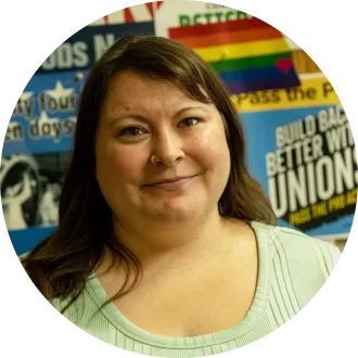 Woman with long hair in green shirt, in front of union posters.