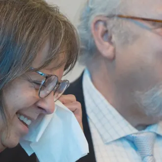 Close-up of man speaking to assembly as woman wipes tear from eye while standing nearby.
